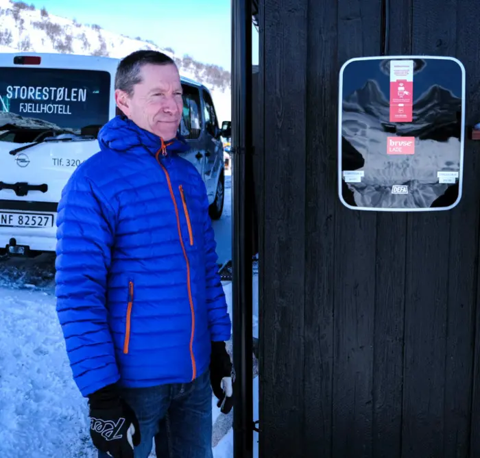 En mann i blå boblejakke står ved siden aven svart ladeboks som er festet på en svart trevegg. I bakgrunnen har du snøfjell, og en hvit bedriftsbil hvor det står