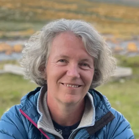 Portrett av en smilende kvinne med kort, grått hår, iført en blå jakke. Bakgrunnen viser et landskap med gress og steiner, og en uskarp, fjern bakgrunn.