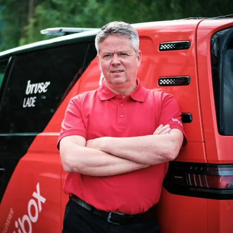 Bildet viser en smilende mann med kort, grått hår, stående med armene i kors, lent mot en svart og rød varebil hvor det står bruse på bilen. Han er iført en rød overdel og svart bukse.