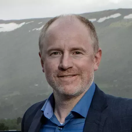Portrett av en smilende mann med kort hår og skjeggstubb, iført en blå dressjakke og skjorte. Bakgrunnen viser et fjellandskap med snøflekker og skyer.