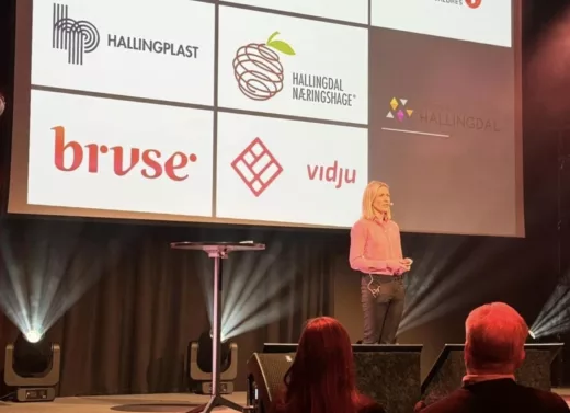 En kvinne står på scenen og taler, mens en projektor i bakgrunnen viser åtte logoer av selskaper. Bilde