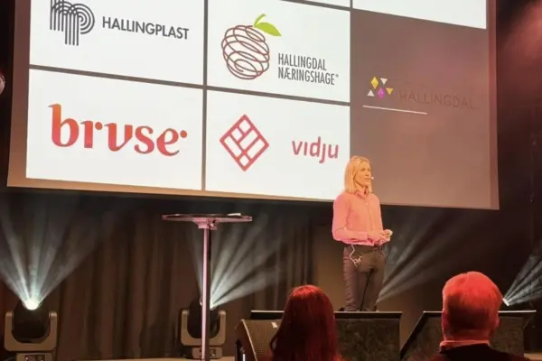 En kvinne står på scenen og taler, mens en projektor i bakgrunnen viser åtte logoer av selskaper. Bilde