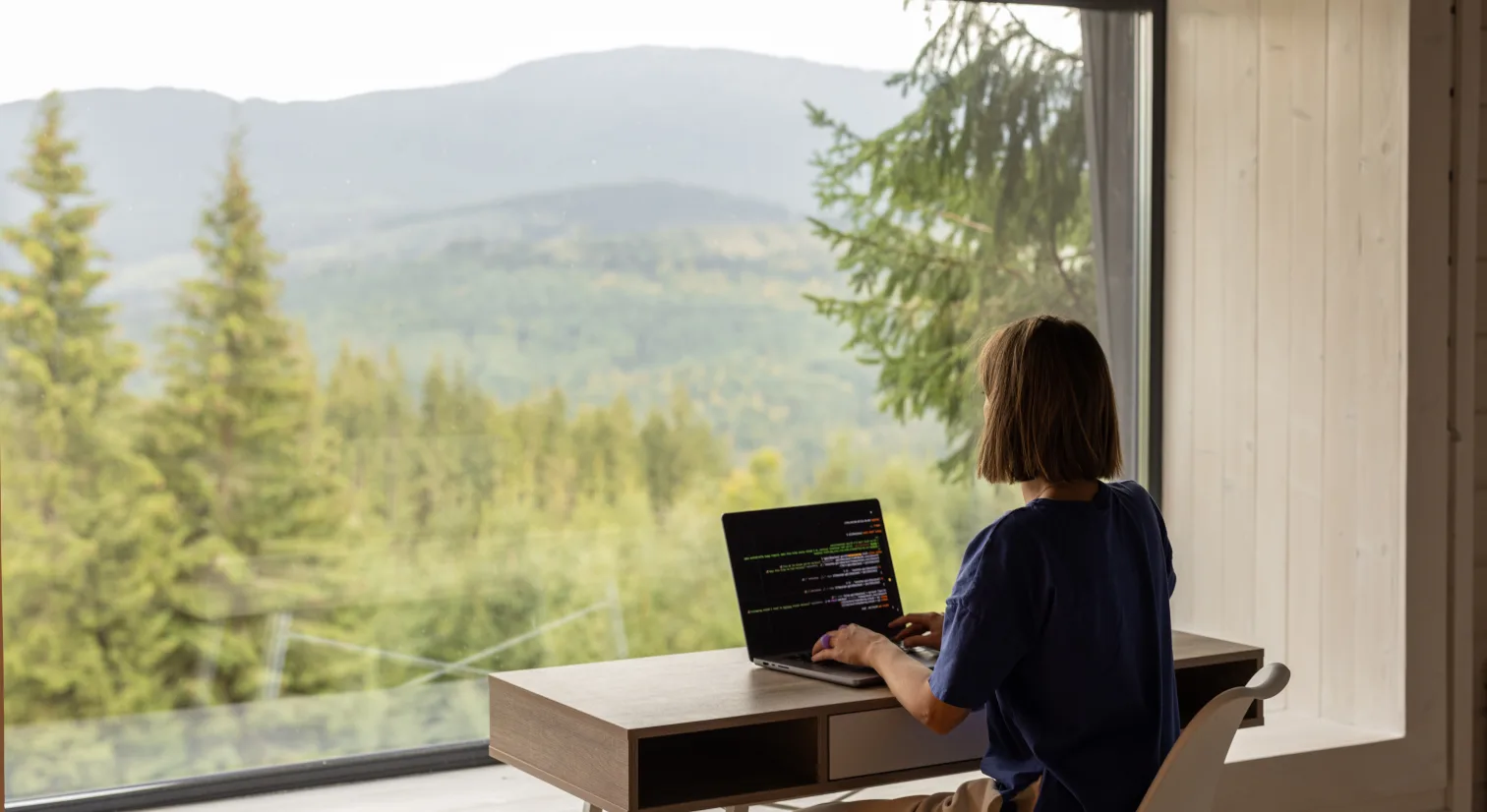 En kvinne sitter ved en pult og skriver på en PC foran et stort vindu som viser en utsikt over en grønn barskog.