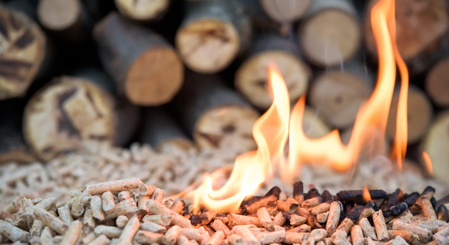 Nærbilde av trepellets med flammer og trekubber i bakgrunnen.
