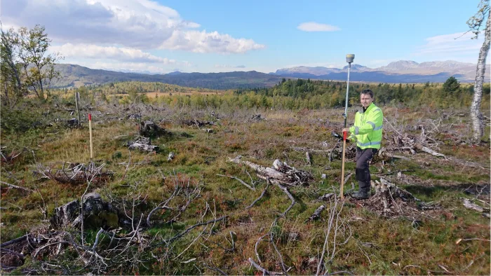 En mann som jobber i Halling Konsult står i en myr med skog og fjell i bakgrunnen, og arbeider med landmålings-apparater.  Han ser inn i kameraet og smiler.